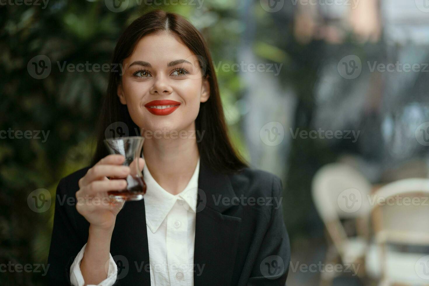 Business brunette woman with red lips smile with teeth drinks tea in a cafe from a Turkish glass mug in a white shirt and black fashion jacket on the street, summer travel, vacation in the city photo