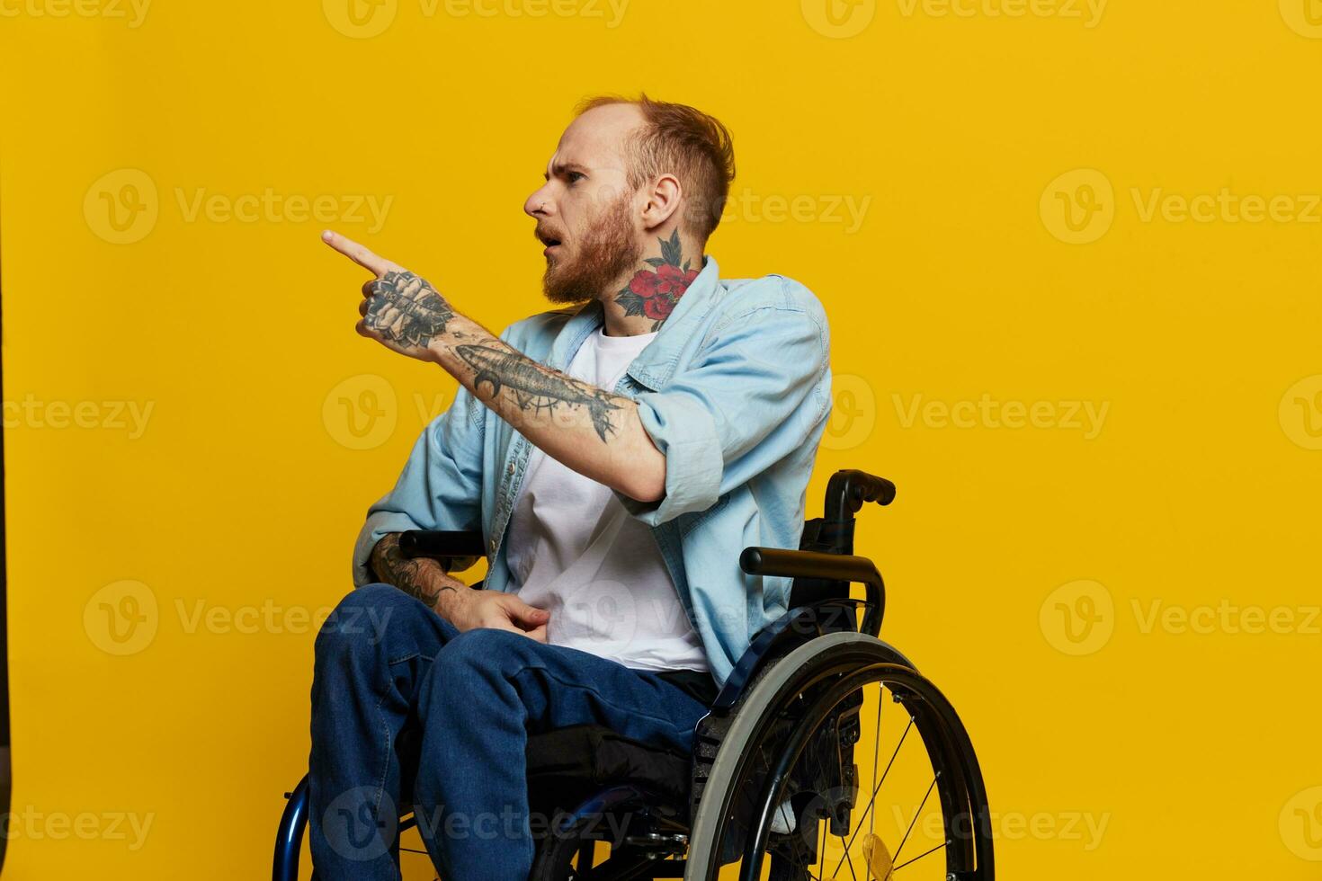 A man in a wheelchair problems with the musculoskeletal system looks at the camera shows a finger on, with tattoos on his hands sits on a yellow studio background, health concept man with disabilities photo