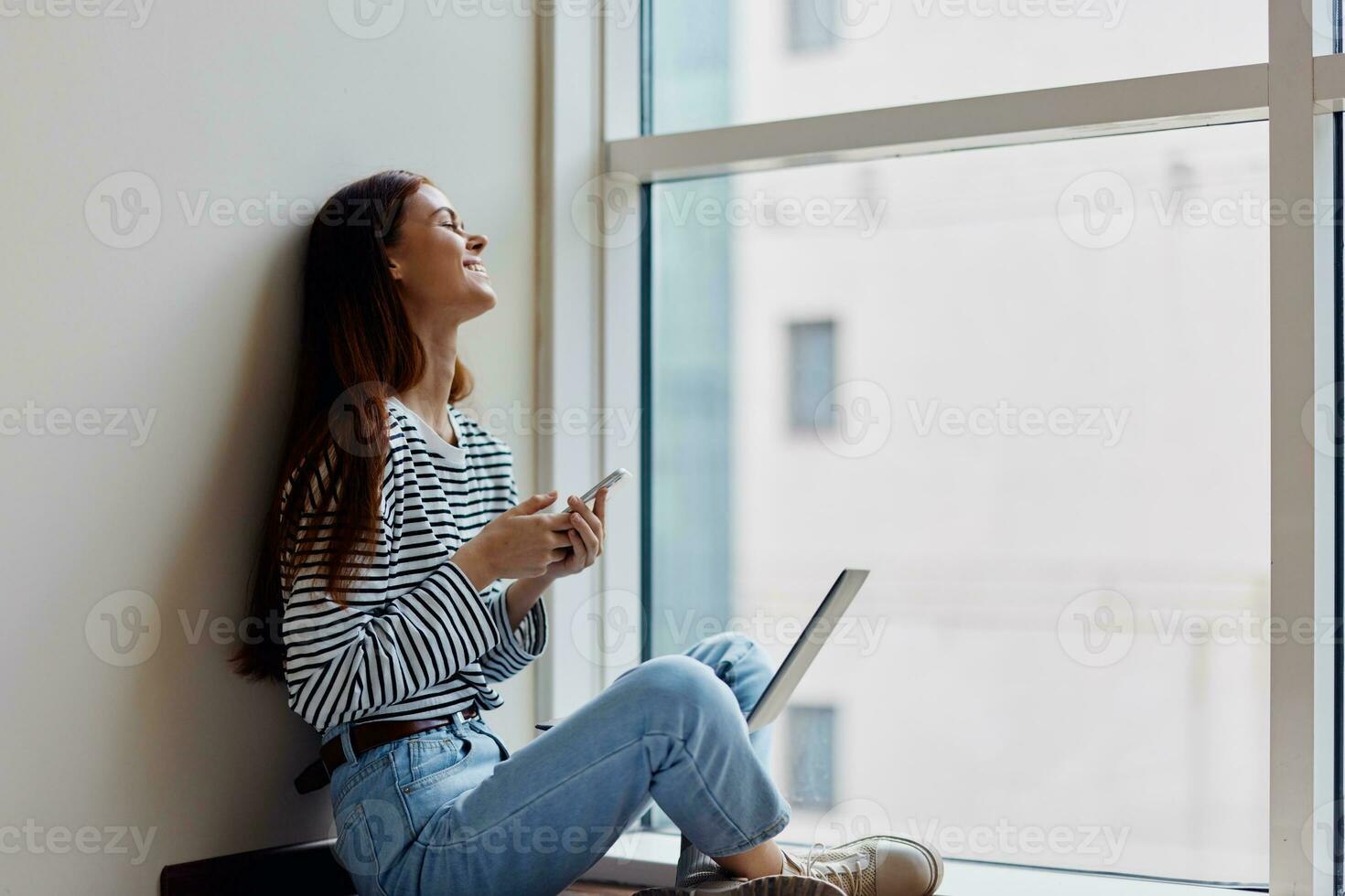 Happy woman sitting on the floor by the windows with a laptop on her lap and holding her phone, a view of the big city, the concept of work and learning online photo