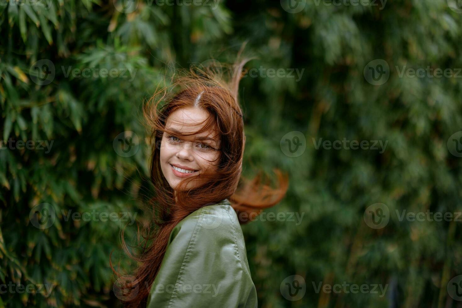 retrato de un hermosa joven niña en el ciudad con un hermosa sonrisa y rojo pelo en un verde impermeable en el ciudad en contra un antecedentes de bambú en primavera, estilo de vida en el ciudad foto