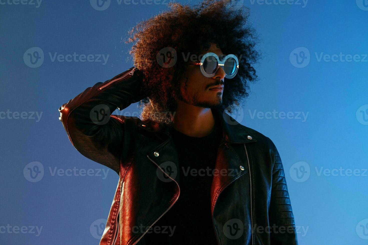 Portrait of a stylish man with curly hair with glasses smile on blue background multinational, color light, black leather jacket trend, modern concept. photo