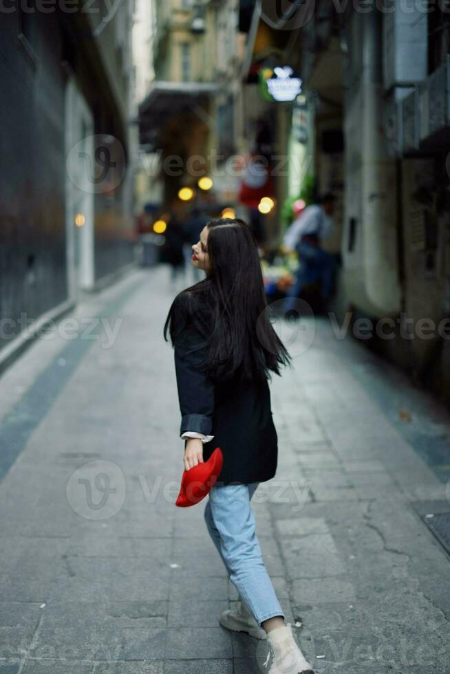 Moda mujer turista en elegante ropa en un chaqueta y rojo boina corriendo abajo el estrecho calle de el ciudad sonrisa y felicidad viaje, francés estilo, cinematográfico color. foto