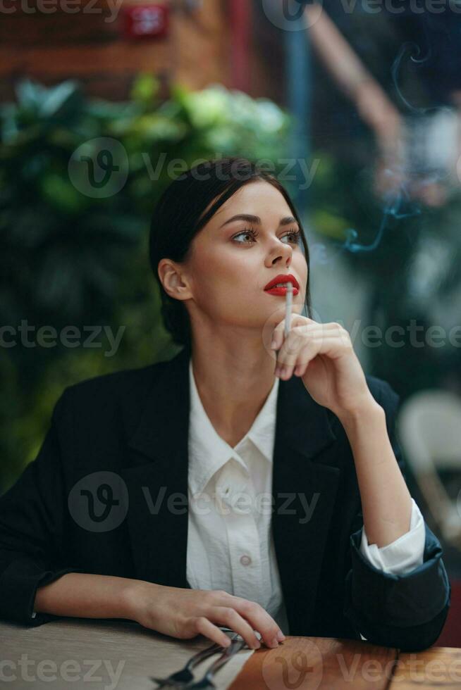 Stylish fashion woman portrait sitting in a cafe at a table and smoking a cigarette releasing smoke from her mouth with red lipstick, a bad habit, a smile with teeth and a thoughtful look photo