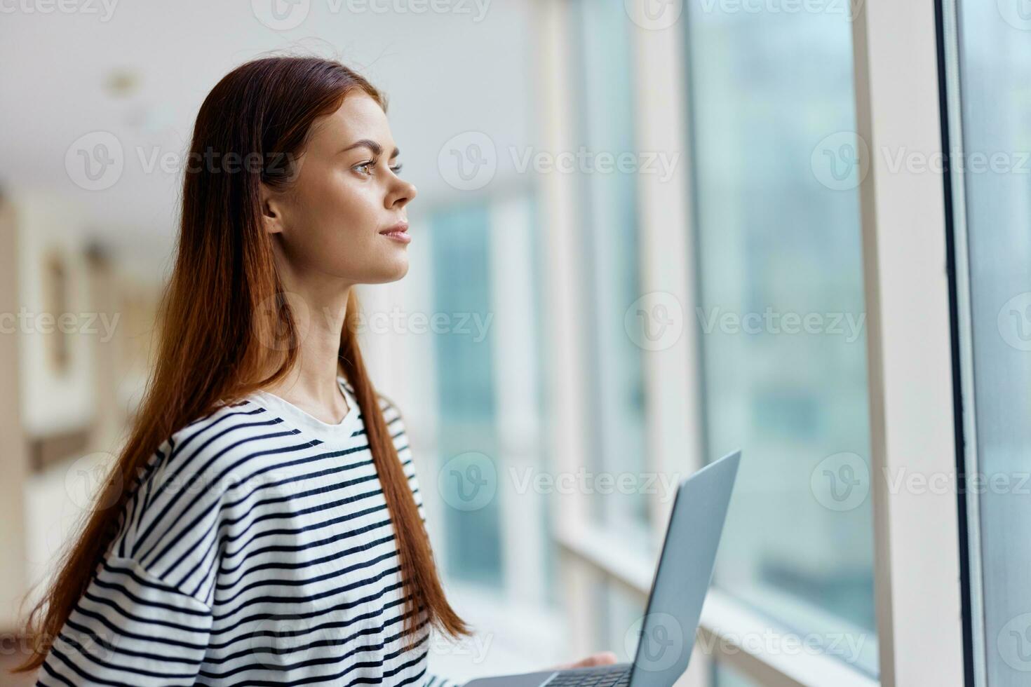 A woman with a laptop in her hands stands near a window in the office at work, a business woman working in a big city, a copy place photo