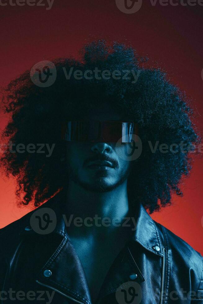 Portrait of fashion man with curly hair on red background with stylish glasses, multicultural, colored light, black leather jacket trend, modern concept. photo