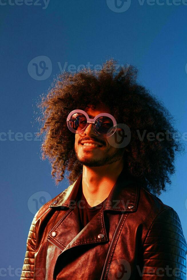 Portrait of a stylish man with curly hair with glasses smile on blue background multinational, color light, black leather jacket trend, modern concept. photo