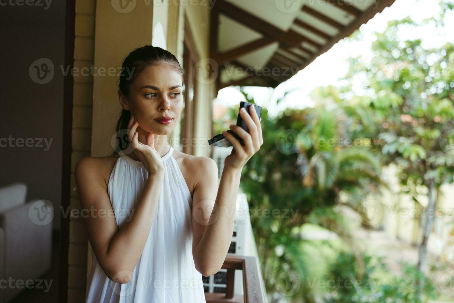 A young beautiful woman looks at her face with makeup in the mirror outside in the summer, protective spf products for problem skin with acne. photo