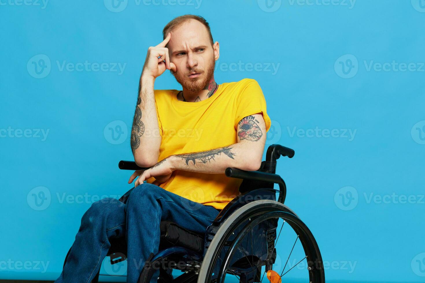 a man sits in a wheelchair thoughtfulness in a t-shirt on a blue background in the studio, the concept of a free barrier-free environment for people with disabilities photo
