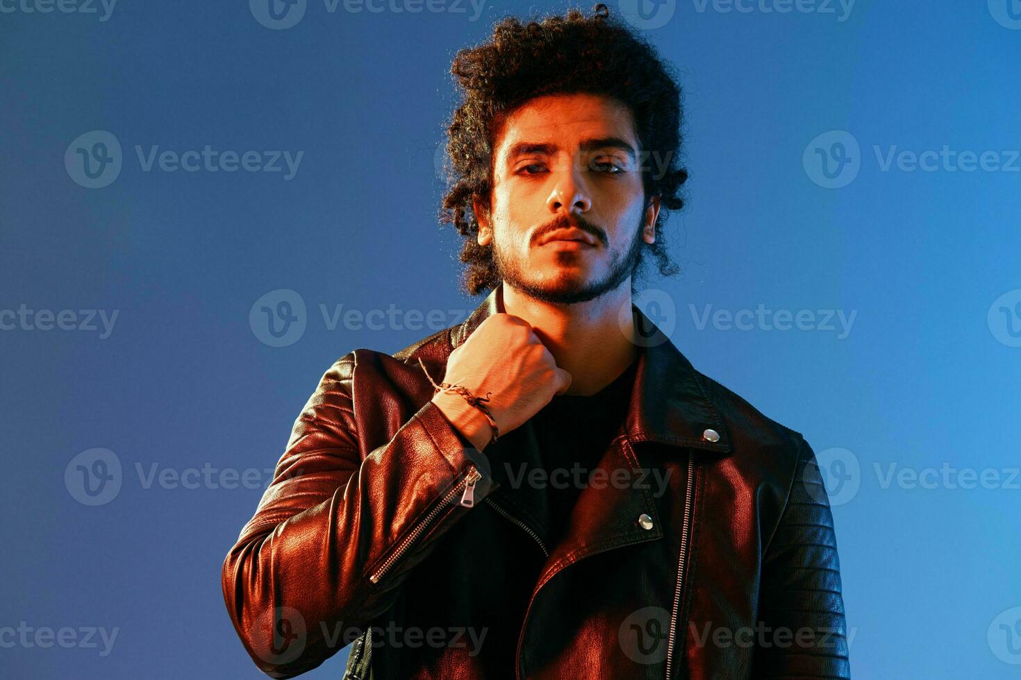 Portrait of a stylish man with curly hair on a blue background multinational, colored light, black leather jacket trend, modern concept. photo