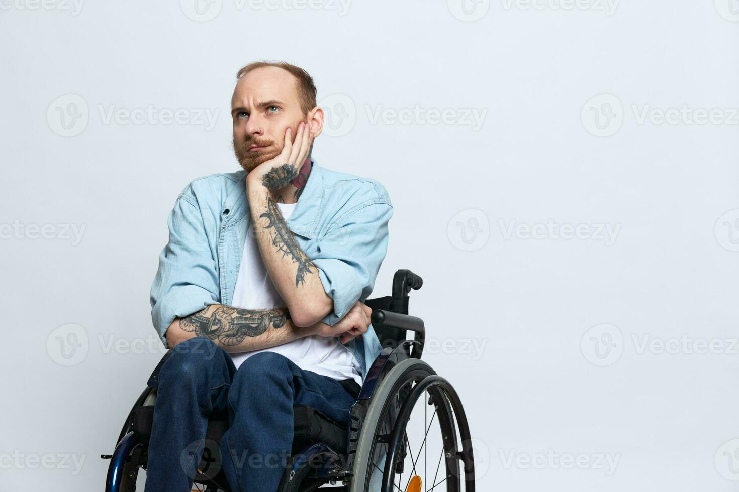 A man in a wheelchair looks at the camera arrogance, with tattoos on his arms sits on a gray studio background, the concept of health is a person with disabilities photo