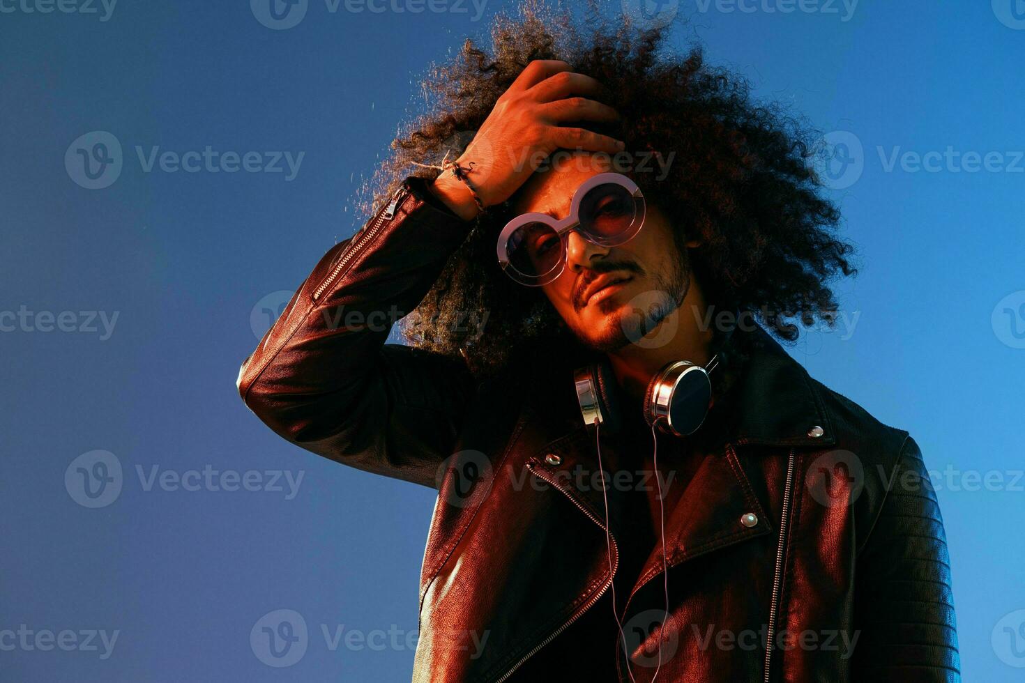 Portrait of a stylish man with curly hair with glasses and headphones on a blue background multinational, colored light, black leather jacket trend, modern concept. photo
