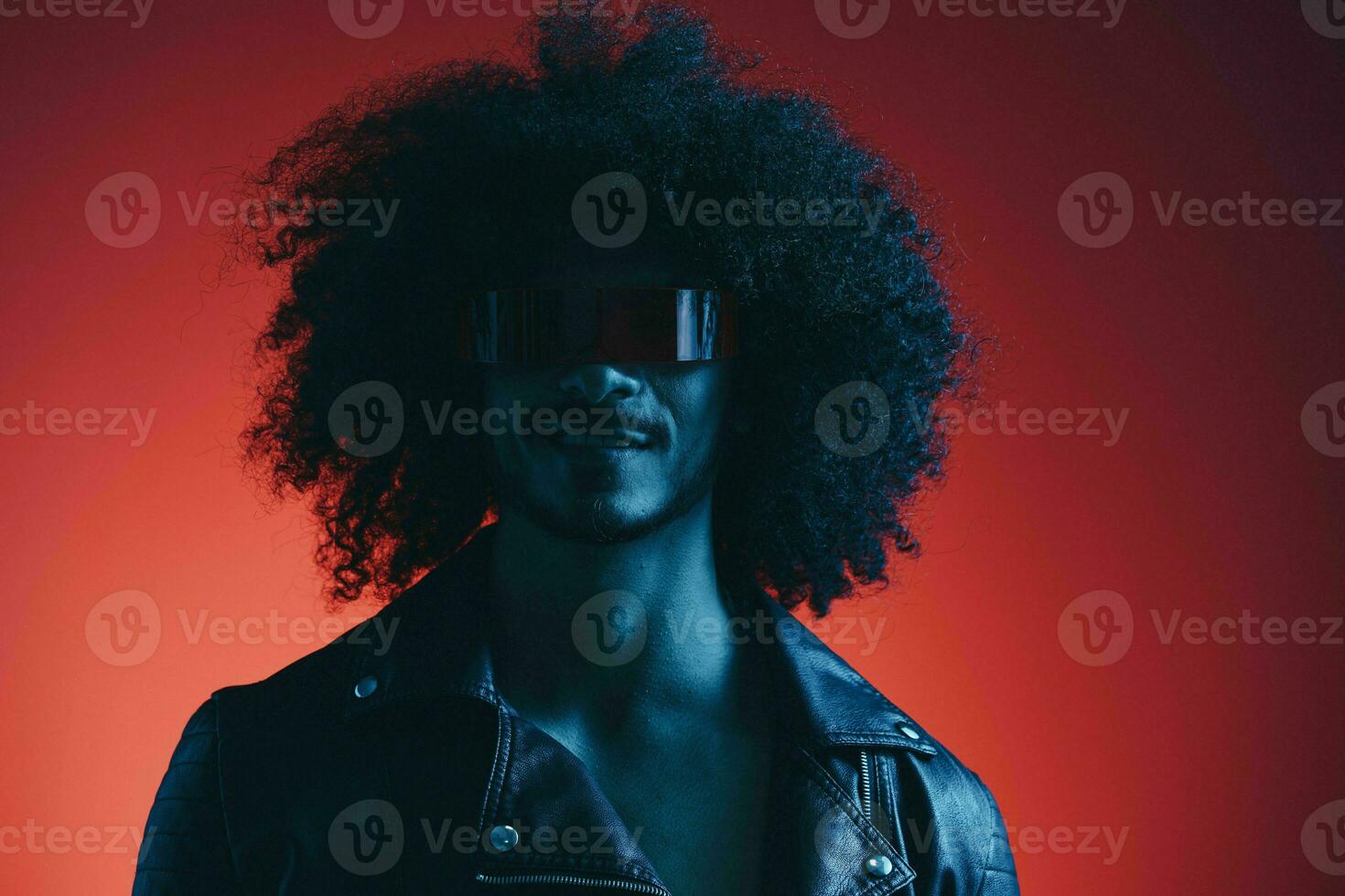 Portrait of fashion man with curly hair on red background with stylish glasses, multicultural, colored light, black leather jacket trend, modern concept. photo