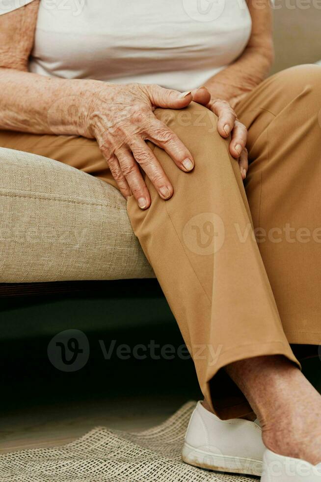 Elderly woman severe pain in her leg sitting on the couch, health problems in old age, poor quality of life. Grandmother with gray hair holds on to her sore knee, problems with joints and ligaments. photo