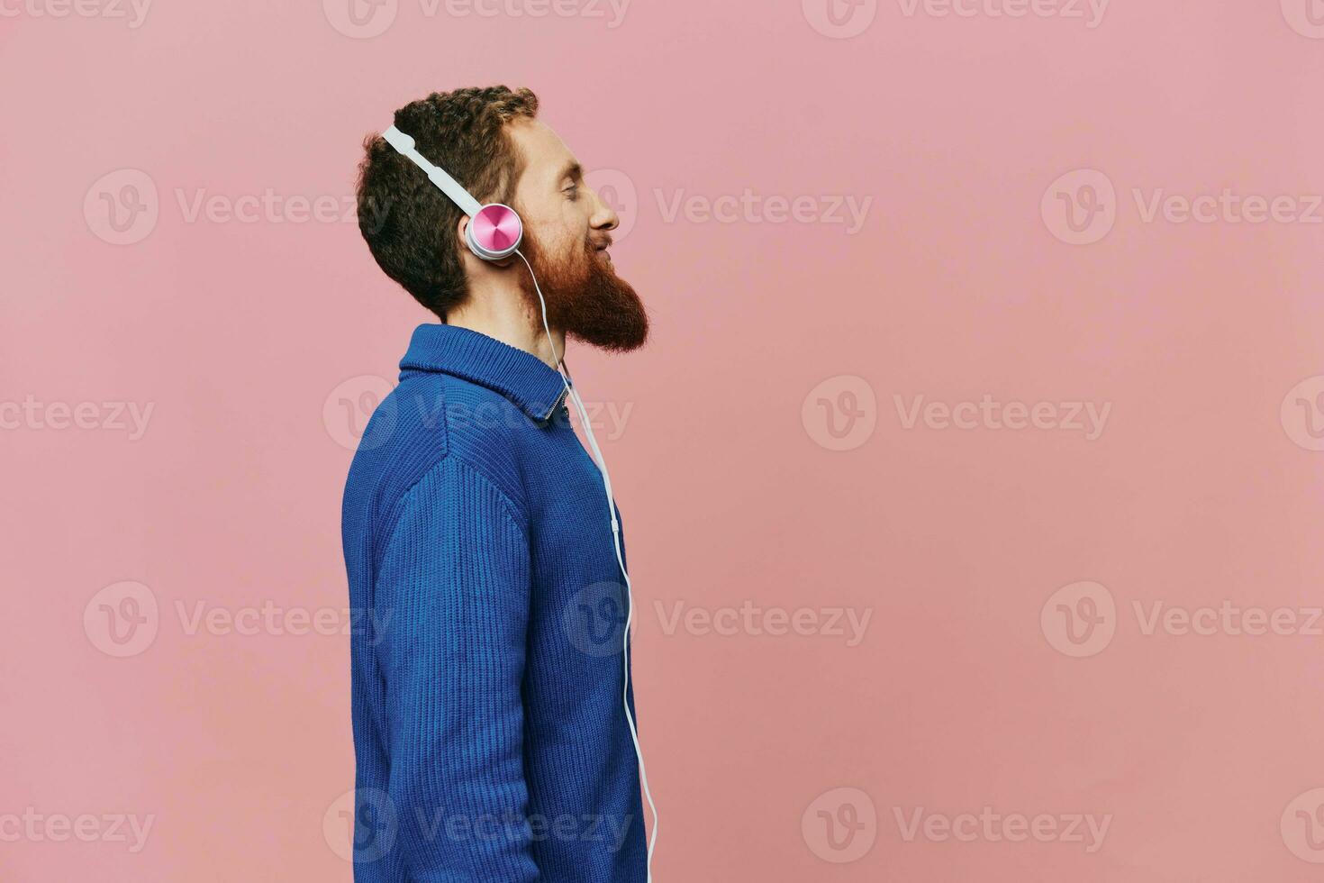 retrato de un pelirrojo hombre vistiendo auriculares sonriente y baile, escuchando a música en un rosado antecedentes. un hipster con un barba. foto