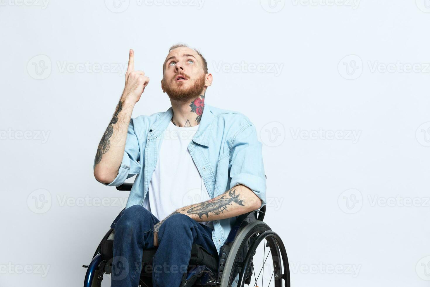 A man in a wheelchair and pointing a finger at, copy space, with tattoos on his hands sits on a gray studio background, health concept a person with disabilities, a real person photo