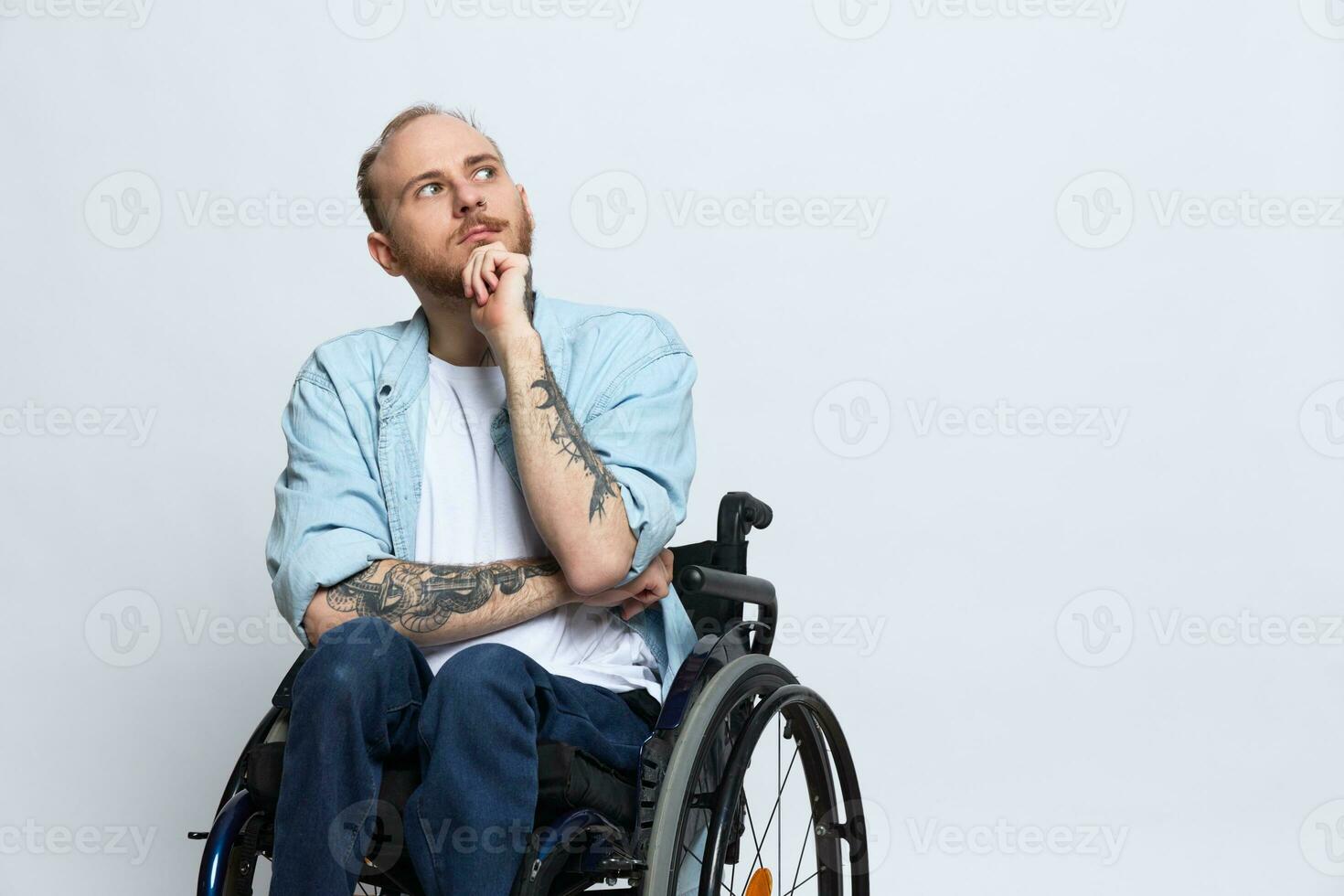 A man in a wheelchair looks at the camera arrogance, with tattoos on his arms sits on a gray studio background, the concept of health is a person with disabilities photo