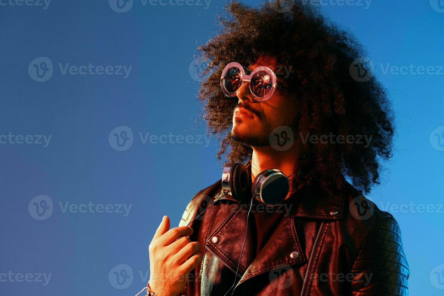Portrait of a stylish man with curly hair with glasses and headphones on a blue background multinational, colored light, black leather jacket trend, modern concept. photo