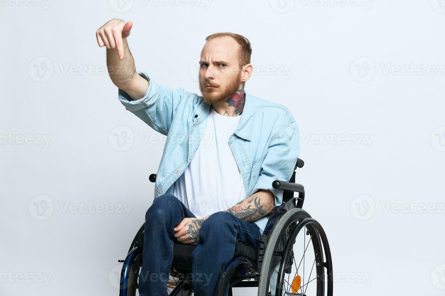 un hombre en un silla de ruedas y señalando un dedo en, Copiar espacio, con tatuajes en su manos se sienta en un gris estudio fondo, salud concepto un persona con discapacidades, un real persona foto