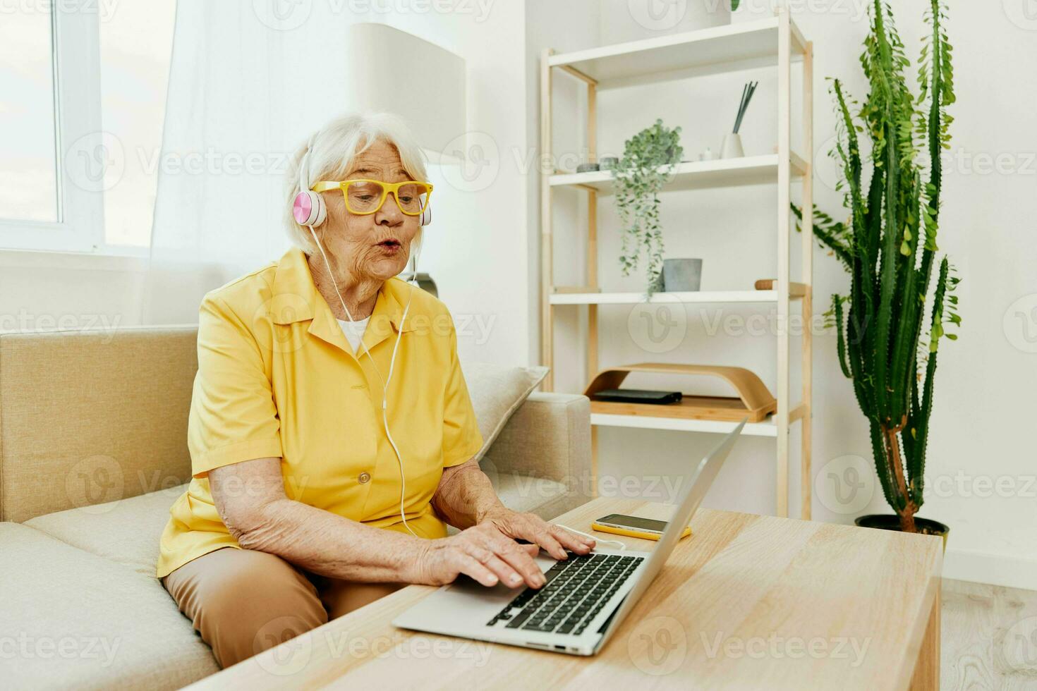 Happy elderly woman with a laptop typing with headphones sitting at home on the couch in a yellow shirt, bright modern interior, lifestyle online communication. photo