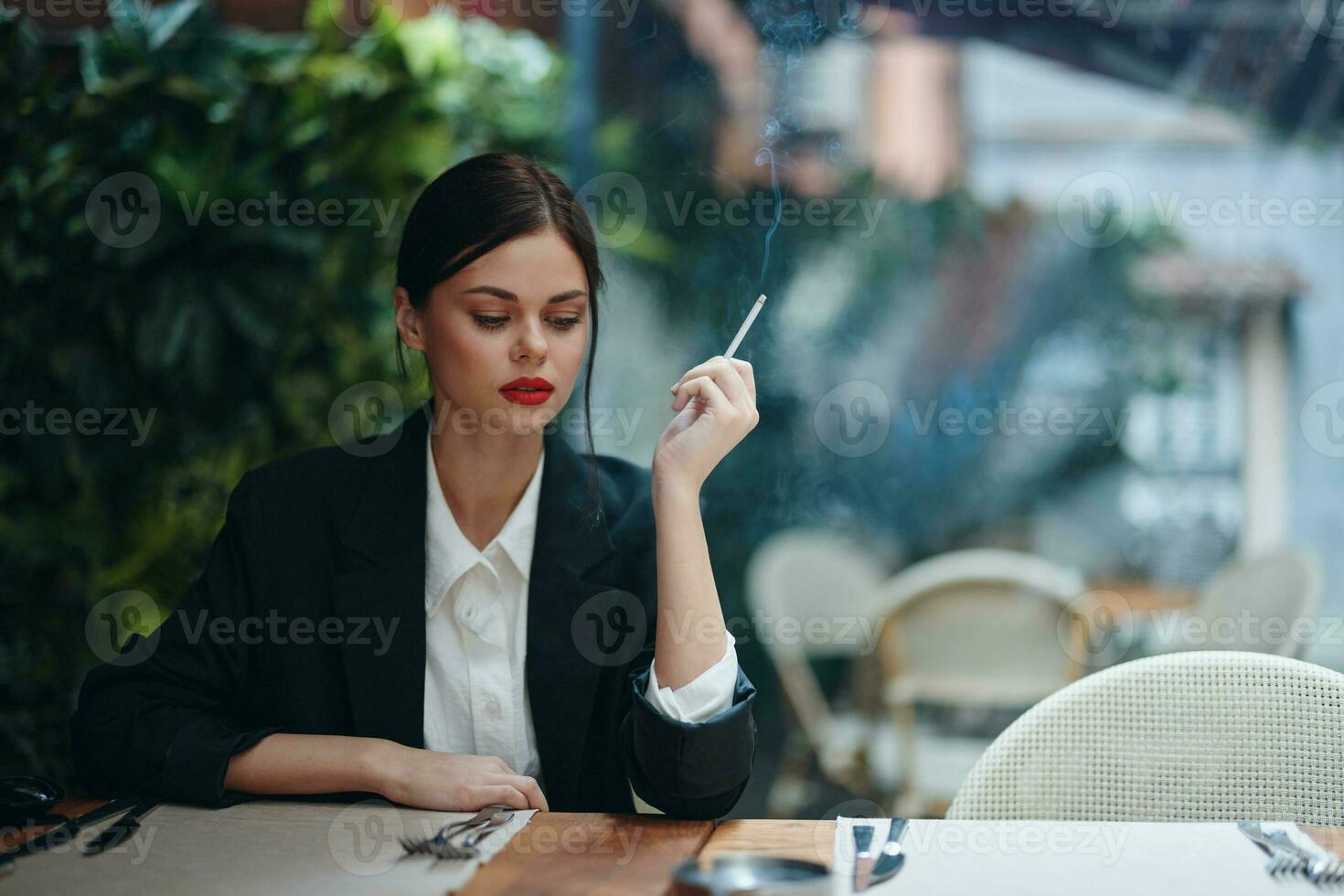 Stylish fashion woman portrait sitting in a cafe at a table and smoking a cigarette releasing smoke from her mouth with red lipstick, bad habit, smile with teeth and pensive look, cinematic vintage photo