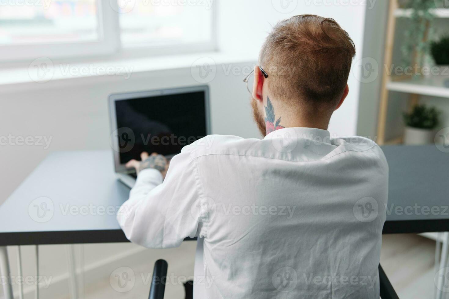 un hombre silla de ruedas empresario con tatuajes oficina trabajos a un ordenador portátil en línea, social redes y puesta en marcha, integración dentro sociedad, el concepto de trabajando un persona con discapacidades, ver desde el espalda foto