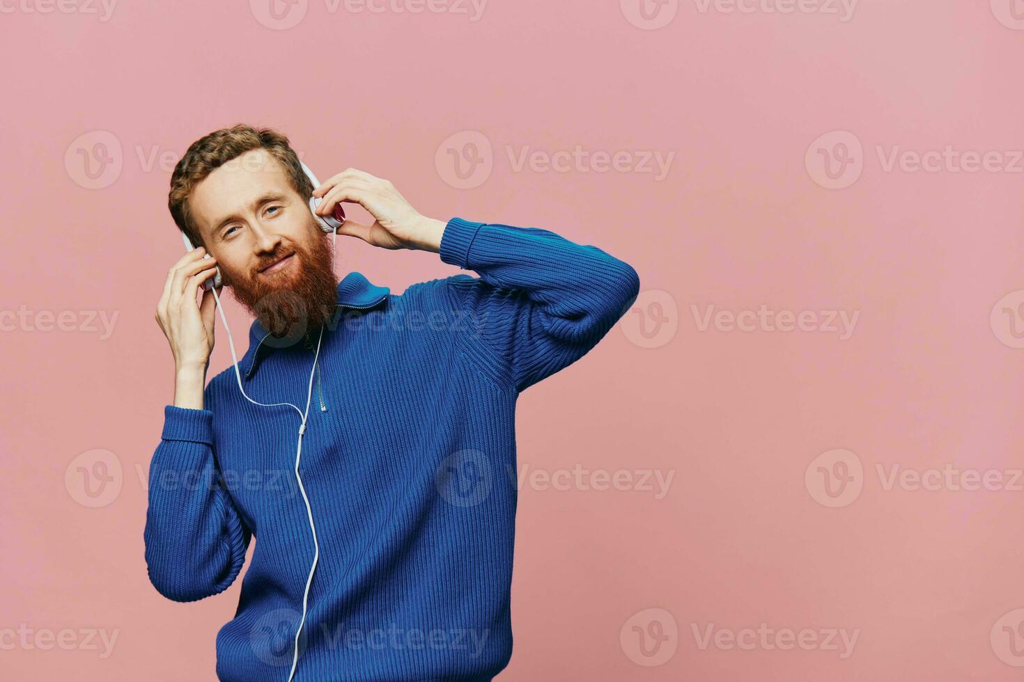 Portrait of a redheaded man wearing headphones smiling and dancing, listening to music on a pink background. A hipster with a beard. photo