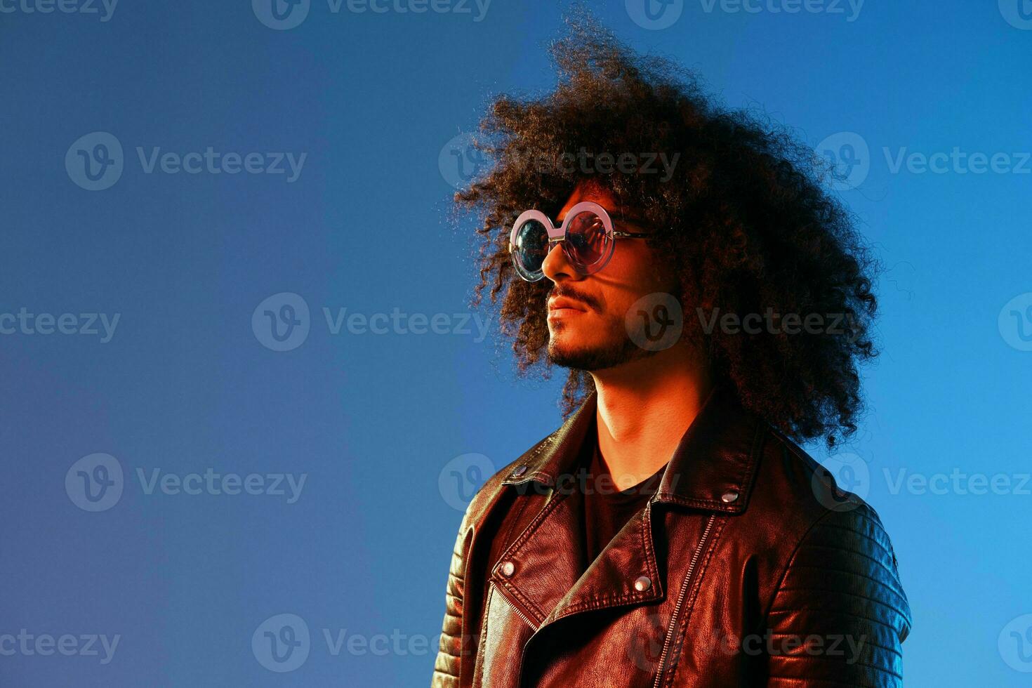 Portrait of a stylish man with curly hair with glasses smile on blue background multinational, color light, black leather jacket trend, modern concept. photo