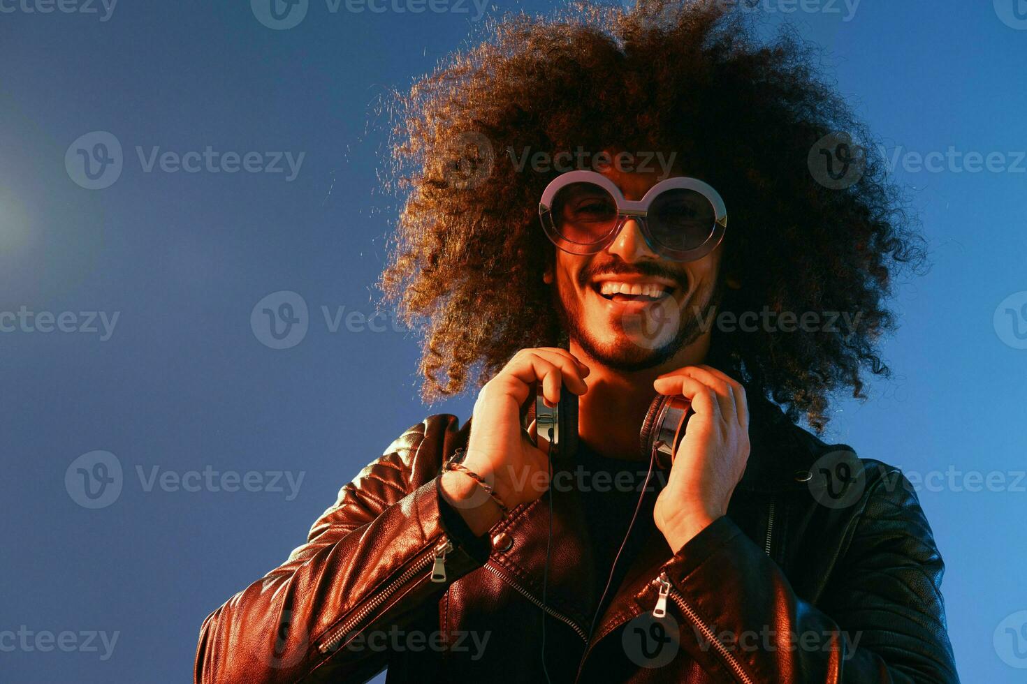 Portrait of a stylish man with curly hair with glasses and headphones on a blue background multinational, colored light, black leather jacket trend, modern concept. photo