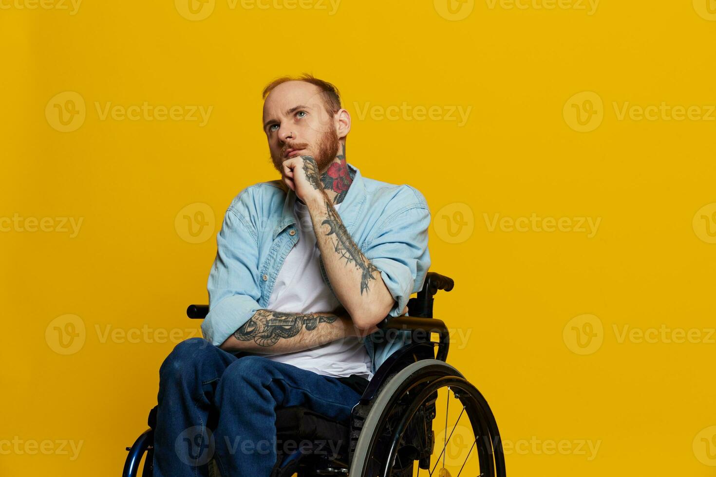 A man in a wheelchair problems with the musculoskeletal system looks at the camera thoughtfully with tattoos on his arms sits on a yellow studio background, health concept a person with disabilities photo