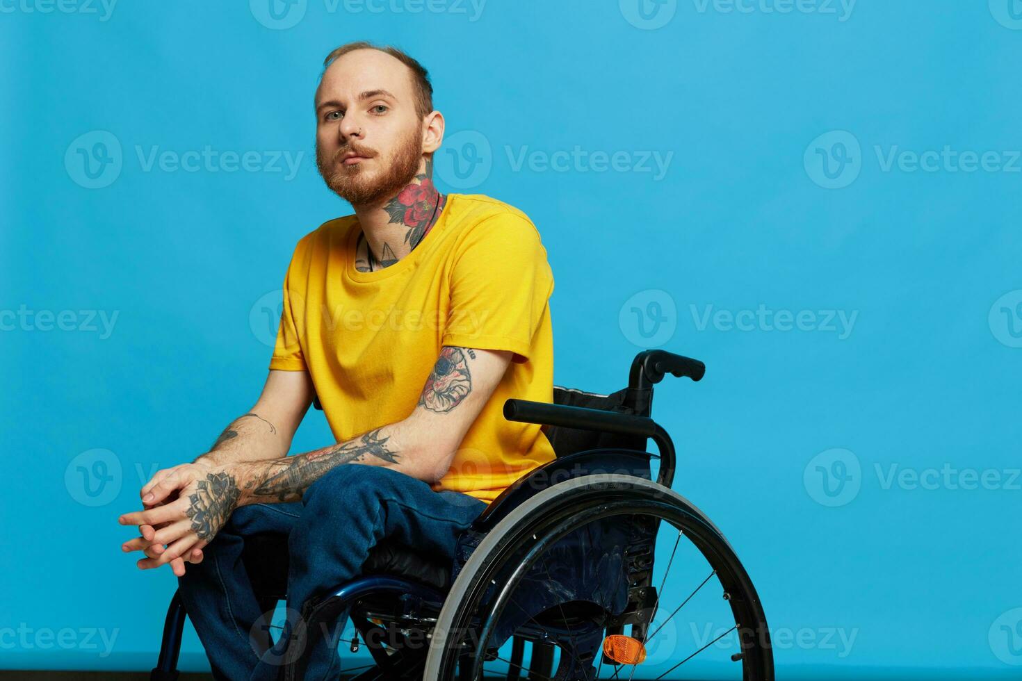 A man in a wheelchair problems with the musculoskeletal system looks at the camera in a t-shirt with tattoos on his arms sits on a blue studio background, full life, real person, health concept photo