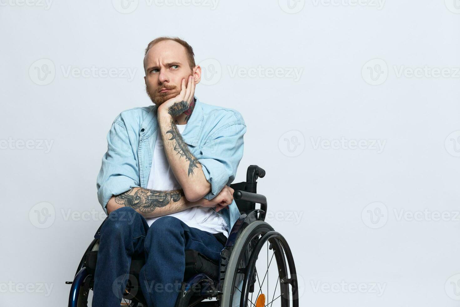 A man in a wheelchair looks at the camera arrogance, with tattoos on his arms sits on a gray studio background, the concept of health is a person with disabilities photo