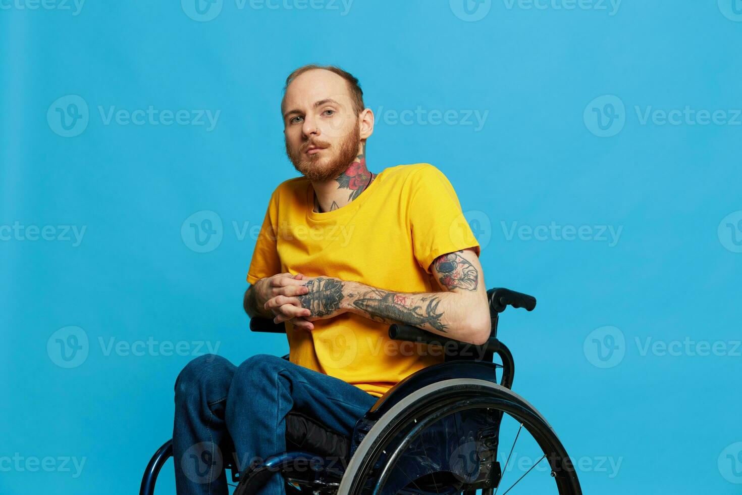 un hombre en un silla de ruedas sonrisa mira a el cámara en un camiseta con tatuajes en su brazos se sienta en un azul estudio fondo, un lleno vida, un real persona foto