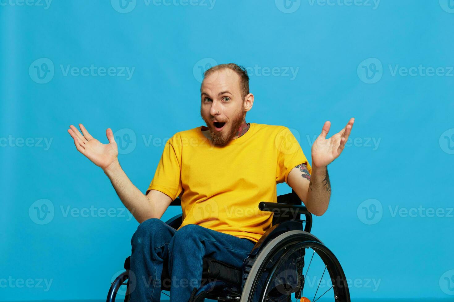 a man sits in a wheelchair thoughtfulness in a T-shirt on a blue background in the studio, Raised his hands up and opened his mouth in surprise, tattooed body photo