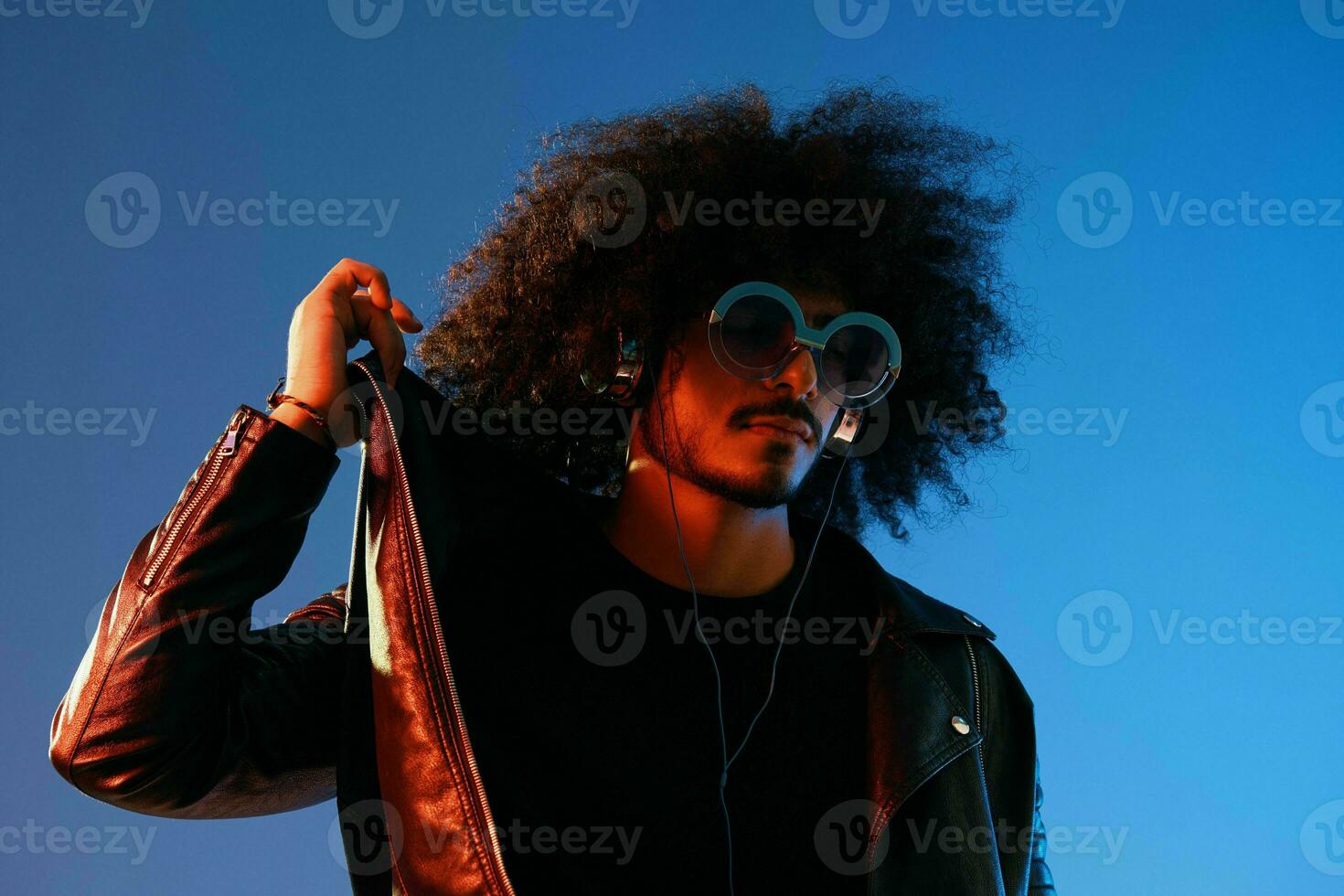 Portrait of a stylish man with curly hair with glasses and headphones on a blue background multinational, colored light, black leather jacket trend, modern concept. photo