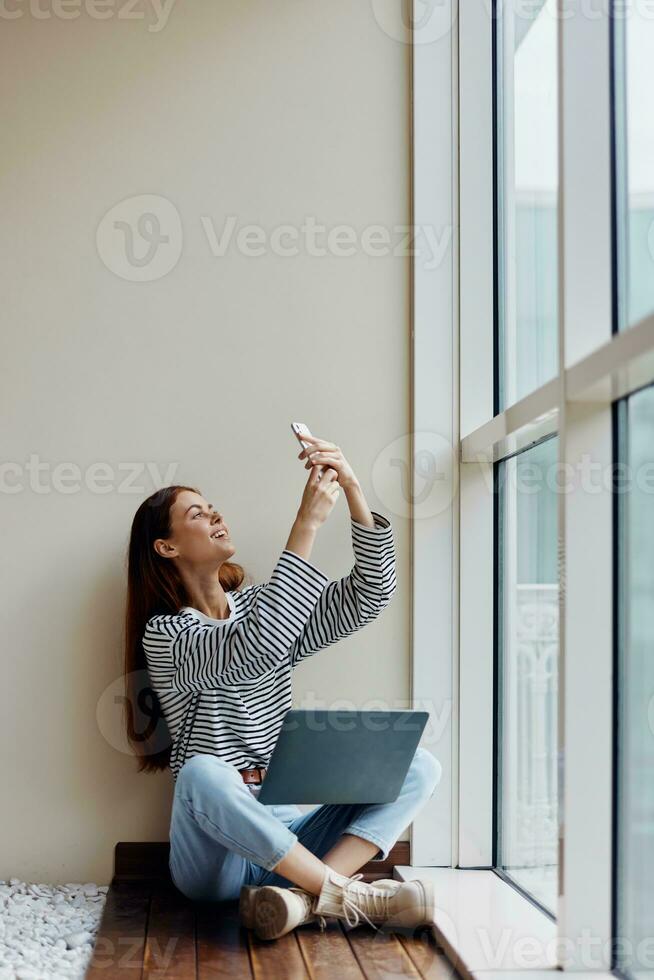 Business woman sitting with laptop and phone at the window full-length and smiling with teeth, happy woman work online in the office, copy space photo
