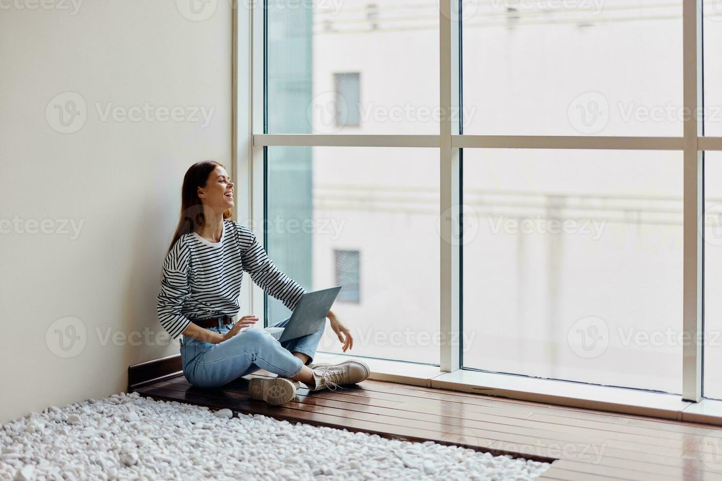 A young woman sits on the floor with her laptop by the window working and studying in the big city photo