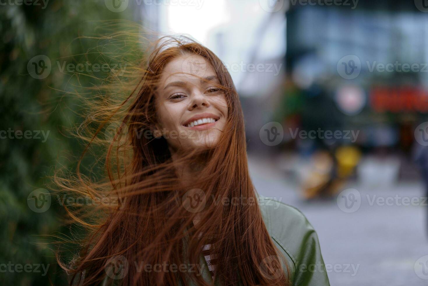 Portrait of a beautiful young girl in the city looking into the camera smile with teeth with red flying hair in a raincoat in the city against a background of bamboo in spring, lifestyle in the city photo
