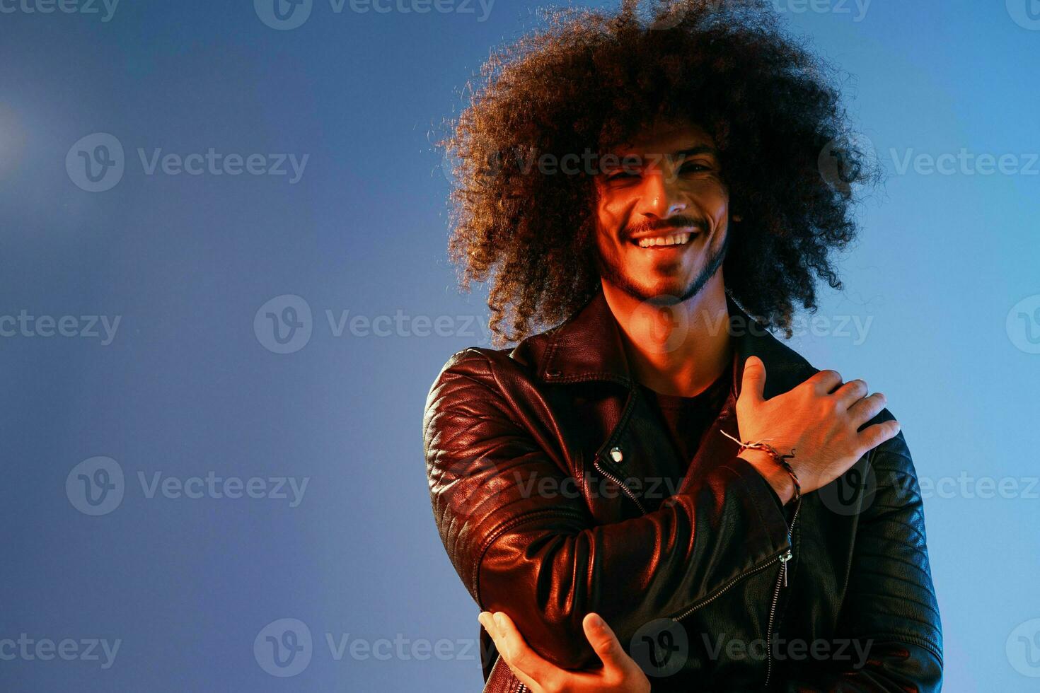 Portrait of a stylish man with curly hair on a blue background multinational, colored light, black leather jacket trend, modern concept. photo