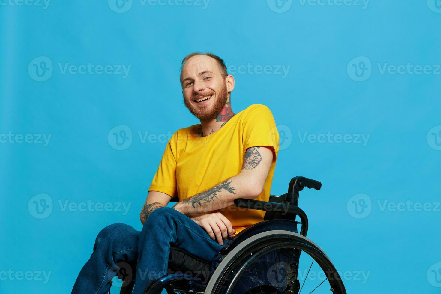 A man in a wheelchair smile looks at the camera in a t-shirt with tattoos on his arms sits on a blue studio background, a full life, a real person photo