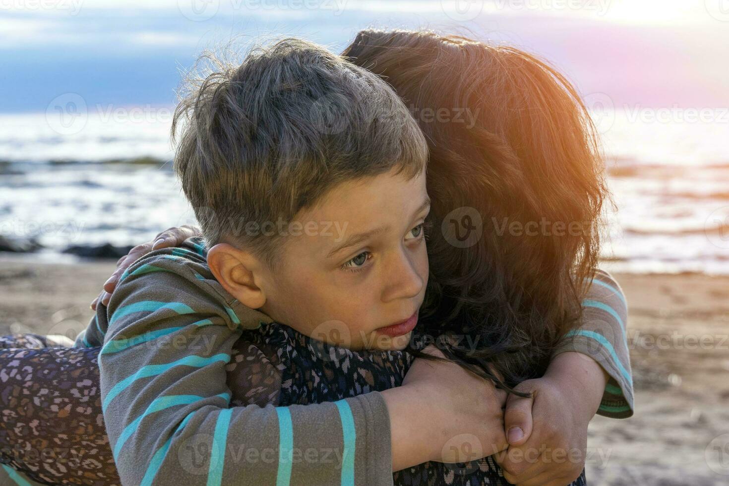 cute sad boy hugs his mother on the seashore in the sunset light photo