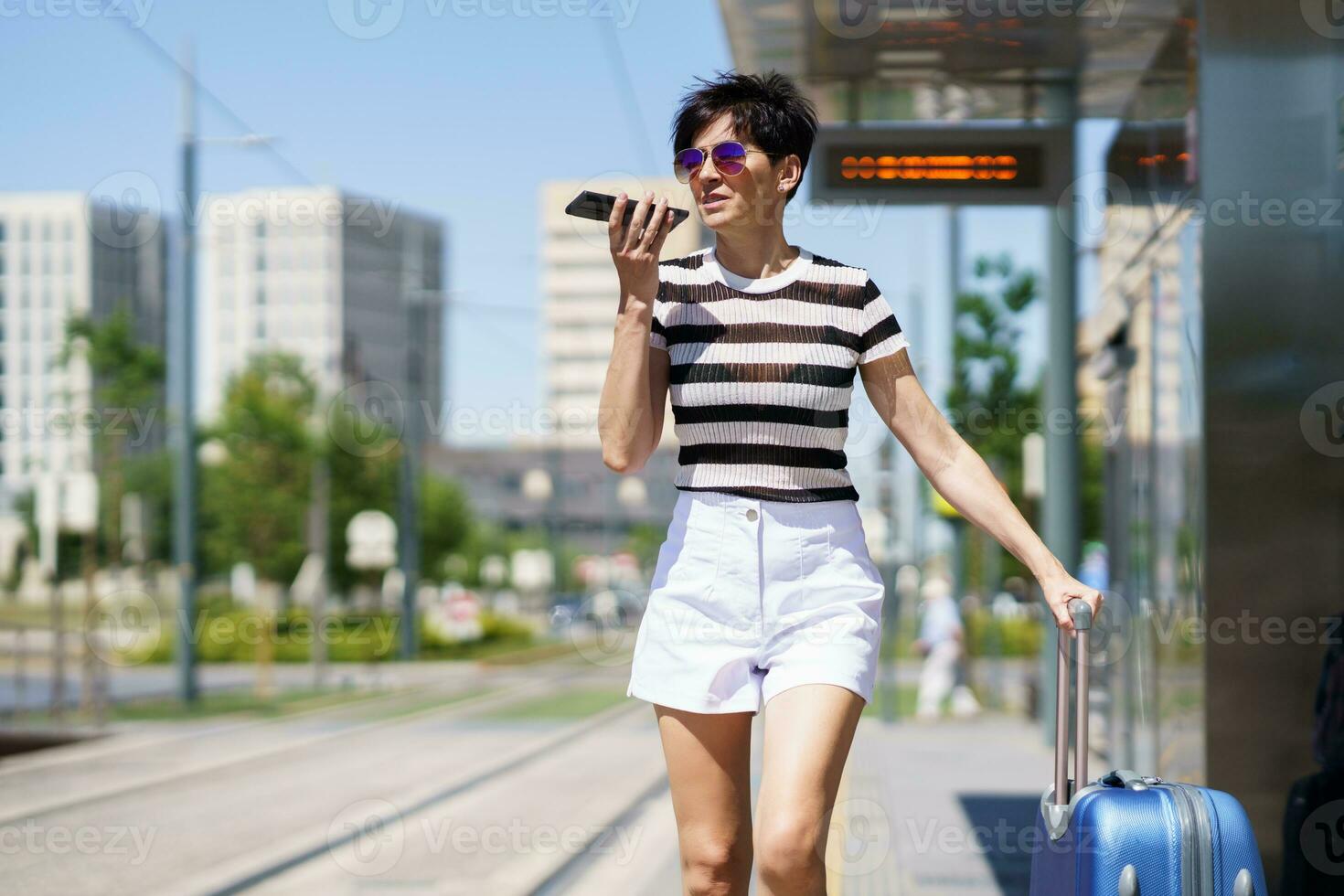 Stylish woman with suitcase recording voice message smartphone in city photo