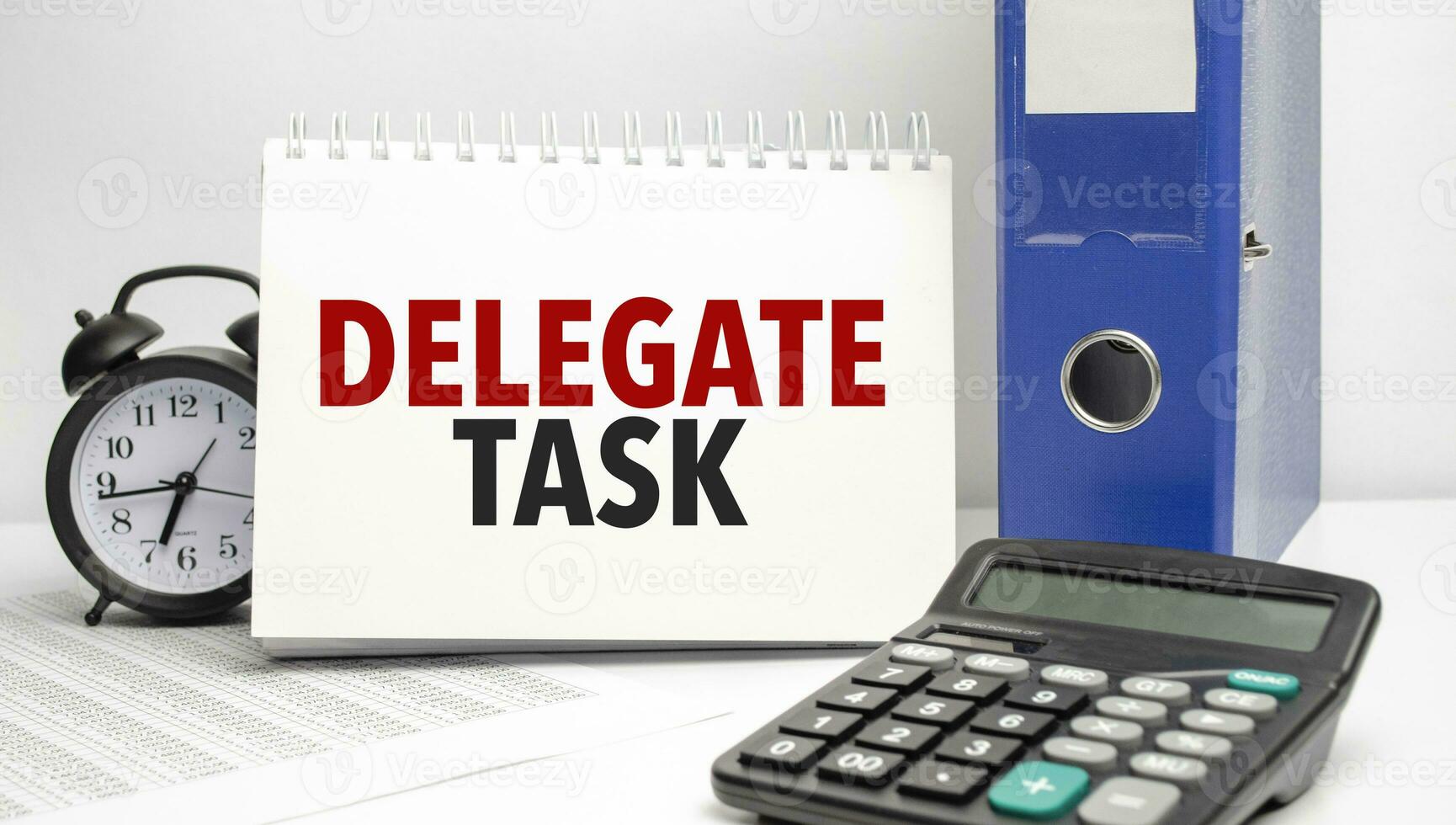delegate task words on white notebook and calculator, black vintage alarm clock and blue paper folder photo