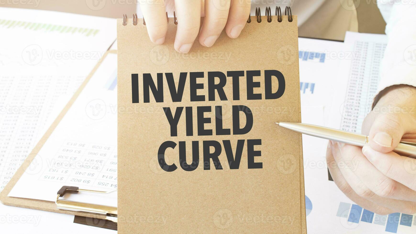 Text INVERTED YIELD CURVE on brown paper notepad in businessman hands on the table with diagram. Business concept photo