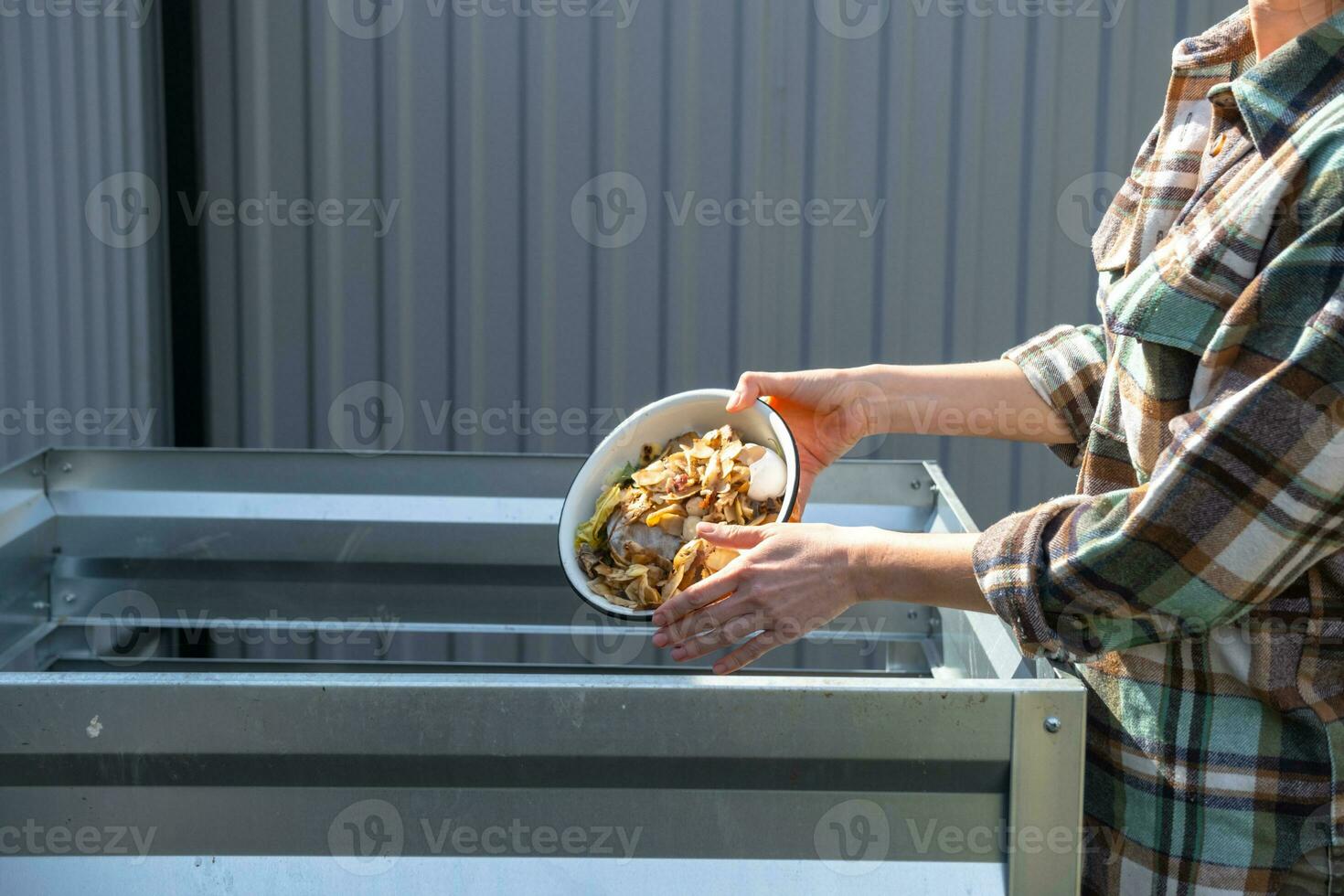 A woman in a plaid shirt pours food waste from a bowl into a compost heap of potato and carrot peelings. Compost box made of metal, eco-friendly fertilizer for the garden photo