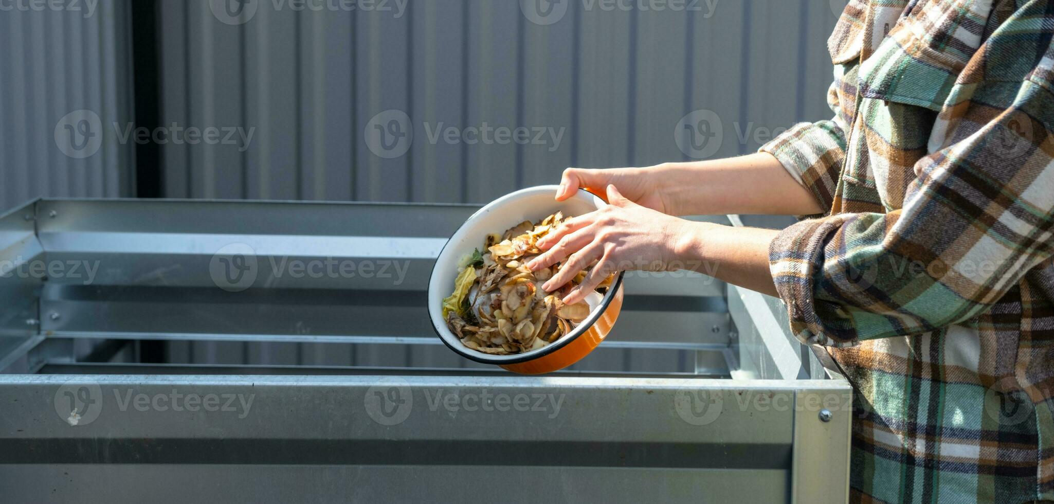 A woman in a plaid shirt pours food waste from a bowl into a compost heap of potato and carrot peelings. Compost box made of metal, eco-friendly fertilizer for the garden photo