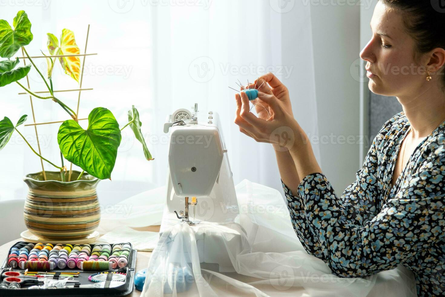 un mujer cose tul en un eléctrico de coser máquina en un blanco moderno interior de un casa con grande ventanas, casa plantas. comodidad en el casa, un ama de casa pasatiempo foto