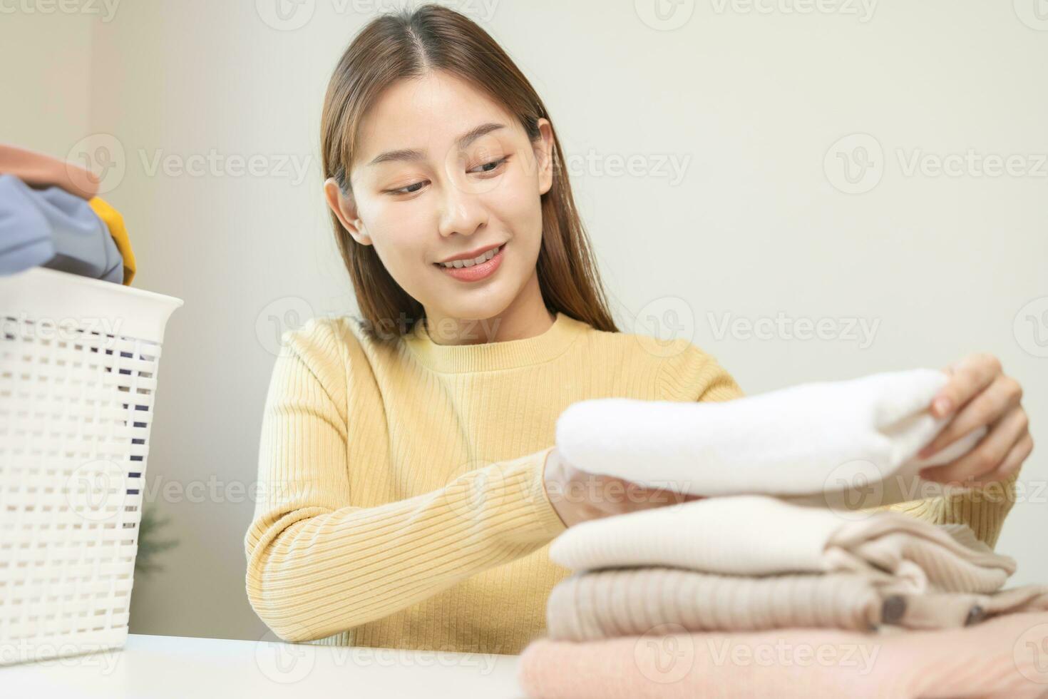 Housewife, asian young woman hand in many folding freshly shirts, sweaters or dress on desk, table after washing clean clothes and drying, making household working in room at home. Laundry and maid. photo