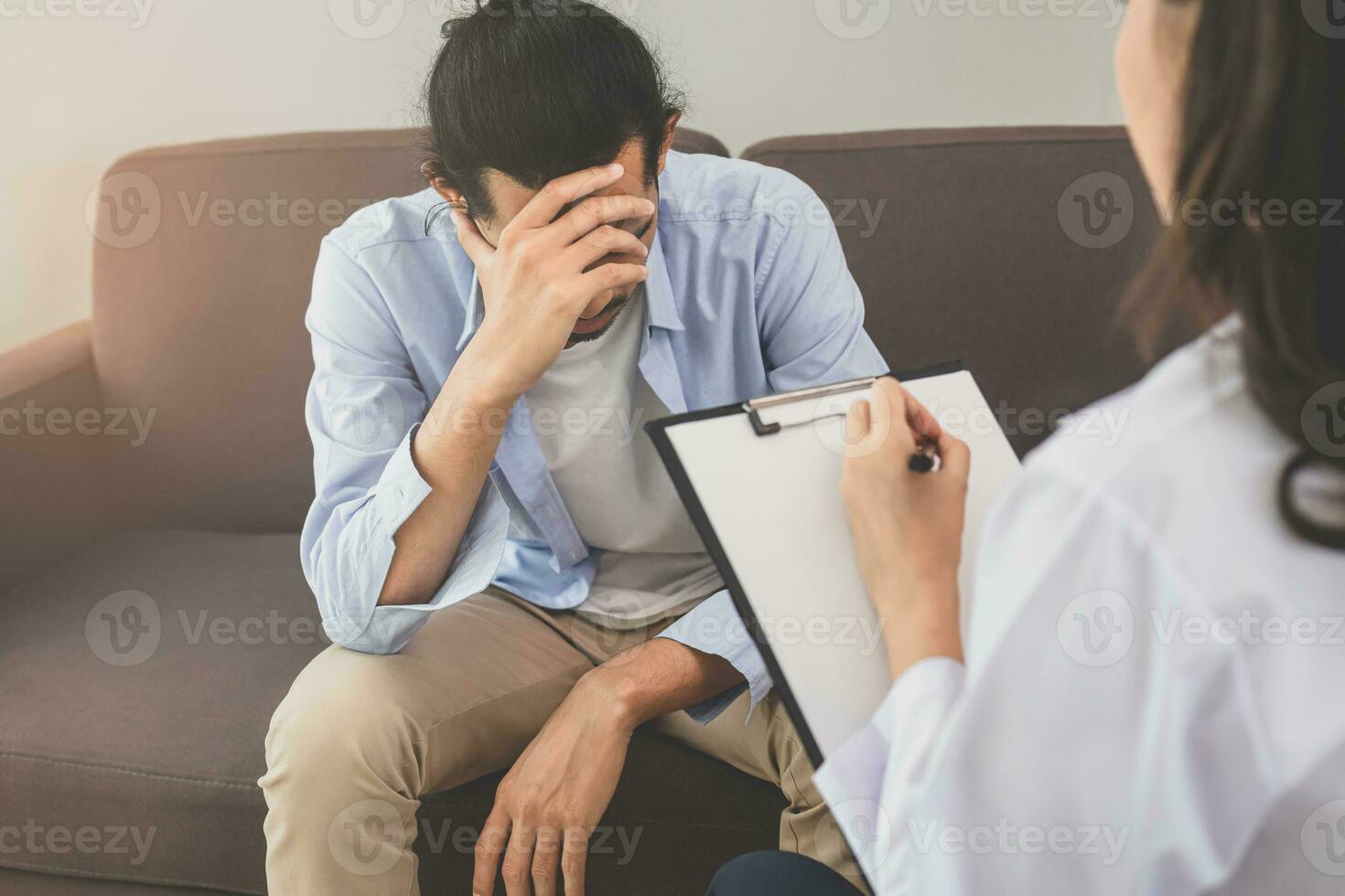 Asian young man, male has mental symptoms which must have been therapy and stress, sitting on couch to consult to psychologist during the session taking notes to find out how to treat the therapist. photo