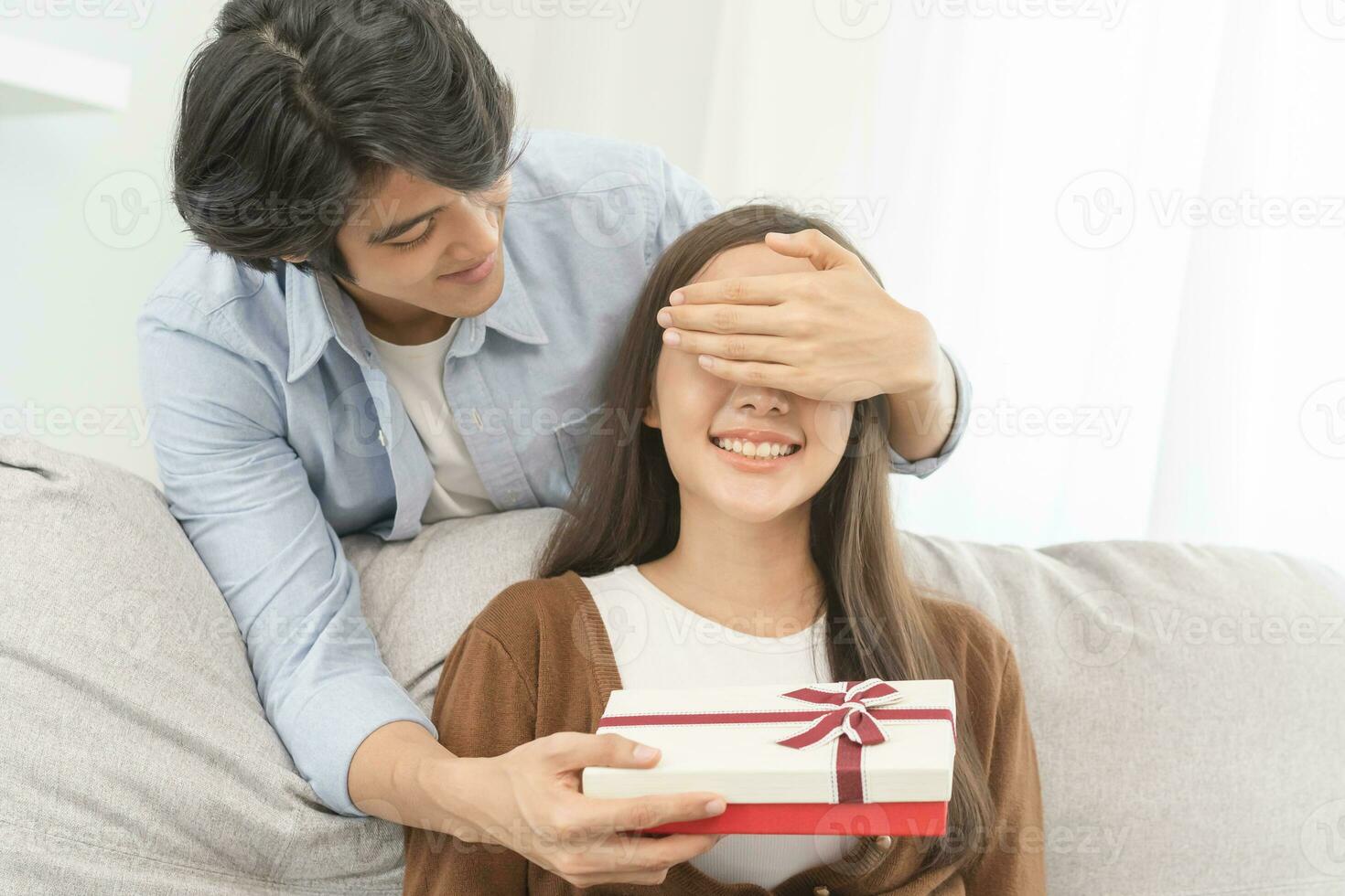 Celebrating anniversary, surprise. Relationship asian young couple love, boyfriend use hand cover girlfriend eye, give a gift by hide box at behind, woman getting present while sitting on sofa at home photo
