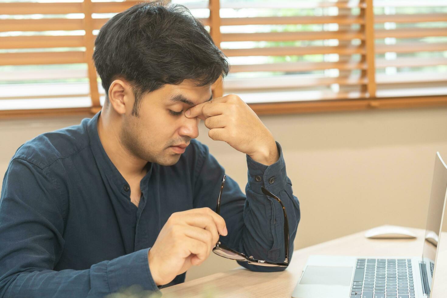 Exhausted, stressed asian young businessman, male working, took off glasses, massage nose from dry eyes, suffer on hard work while use laptop computer at office home. Overtime job, debt problem people photo